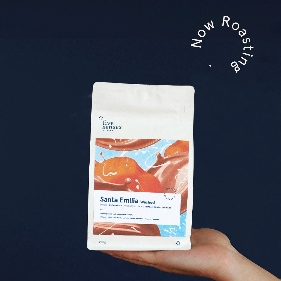 Hand holding a coffee package labeled 'Santa Emilia' against a dark background with 'Now Roasting' text.