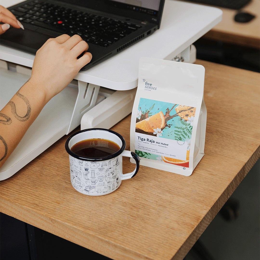 Person using a laptop with a cup of coffee and a bag of tea on a wooden desk.